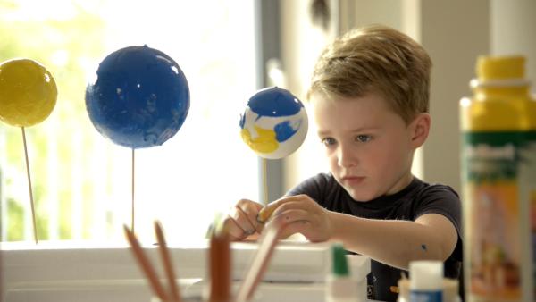 Ein Junge befestigt bunte Planeten aus Styroporkugeln und Holzstäbchen auf einer weißen Unterlage.