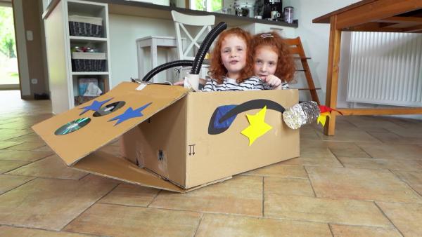 Zwei Mädchen sitzen in ihrem selbst gebastelten Raumschiff aus Karton auf dem Küchenboden.