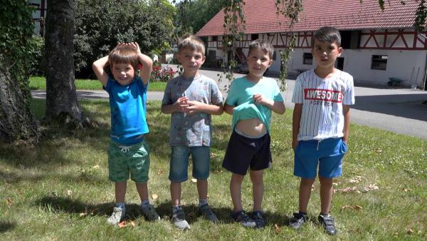 Vier Jungen stehen auf einer Wiese vor einem Bauernhofgebäude. Sie tragen T-Shirts und kurze Hosen.