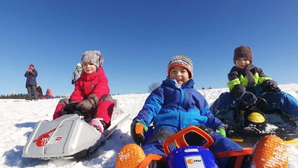 Drei Kinder in warmen Winterklamotten und Mützen sitzen auf Schlitten und lachen in die Kamera.