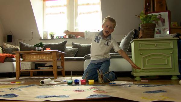 Ein Junge kniet auf dem Boden und lacht. Vor ihm liegen Pinsel, Farben und Papier.