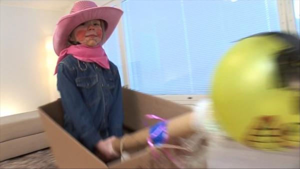 Ein Kind mit einem pinken Westernhut und Jeanshemd in einem Karton. Es hält einen Stab mit einem Ballon in den Händen.
