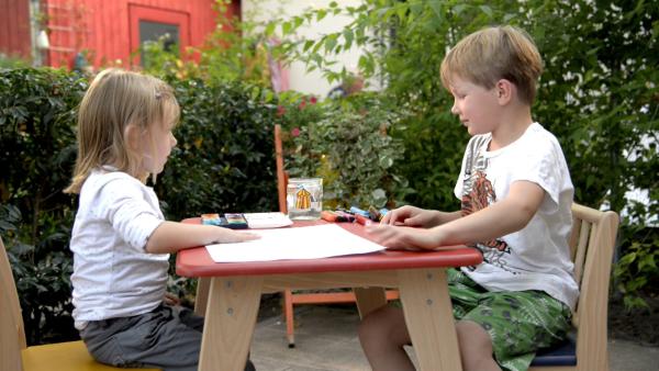 Ein Junge und ein Mädchen sitzen an einem Tisch. Vor dem Mädchen liegt ein weißes Blatt Papier.
