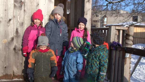 Eine Gruppe Kinder steht vor einer Holzhütte. Sie sind alle warm angezogen, da es geschneit hat.
