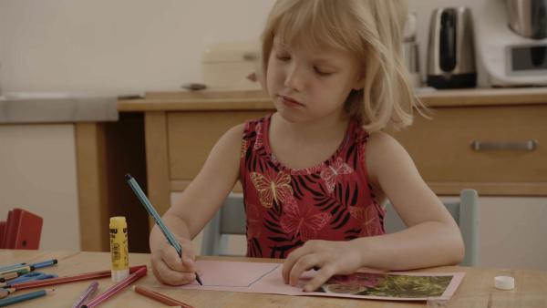 Ein Mädchen sitzt an einem Tisch und hält einen blauen Stift in der Hand. Sie malt auf eine Karte.