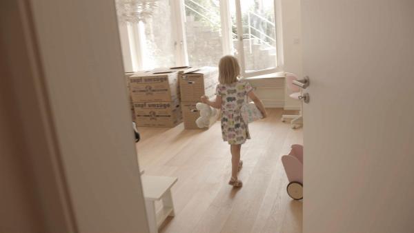 Ein Mädchen läuft mit ihrem Kuscheltier und einer Box in der Hand in ihr neues und leeres Zimmer.