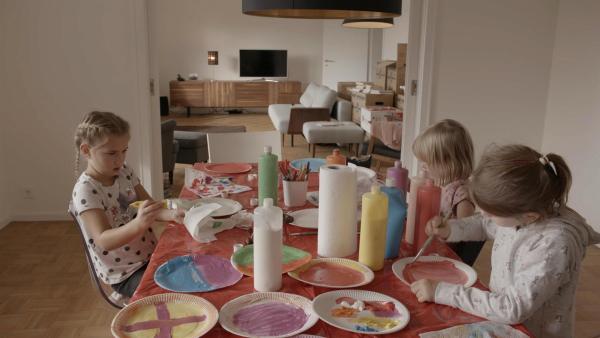Drei Mädchen sitzen an einem Tisch. Auf ihm stehen viele bunte Farben. Damit malen sie Teller an.