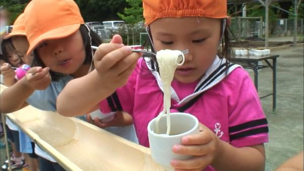 Ein japanisches Kind steht vor einer Wasserrinne. Es hält eine Gabel mit Nudeln und eine Schüssel.