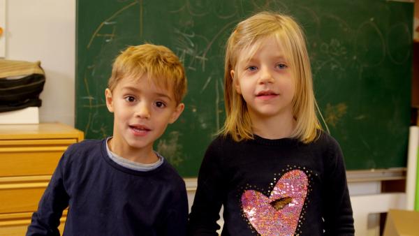 Zwei Kinder stehen vor einer Wand mit einer Tafel und schauen in die Kamera um sich vorzustellen.