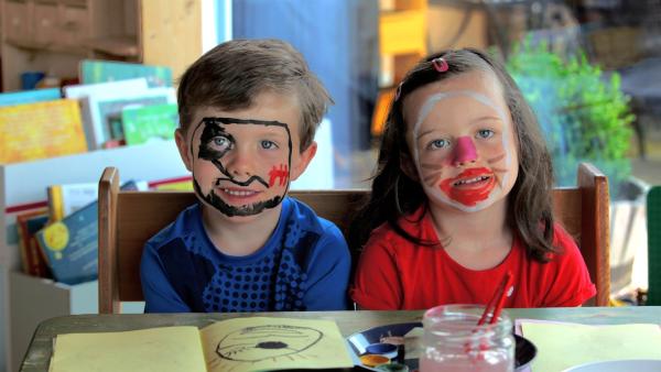 Ein Junge und ein Mädchen sitzen an einem Tisch und malen ein Schminkbuch. Sie sind auch geschminkt.