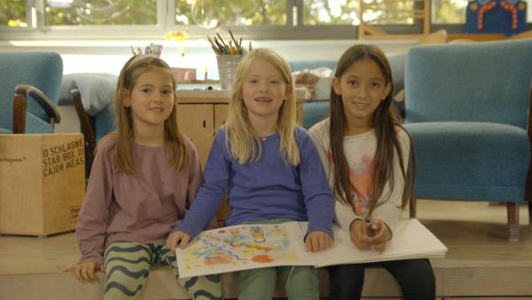 Drei Mädchen sitzen auf einer Couch und halten ein Buch mit selbst gemalten Bildern in der Hand.