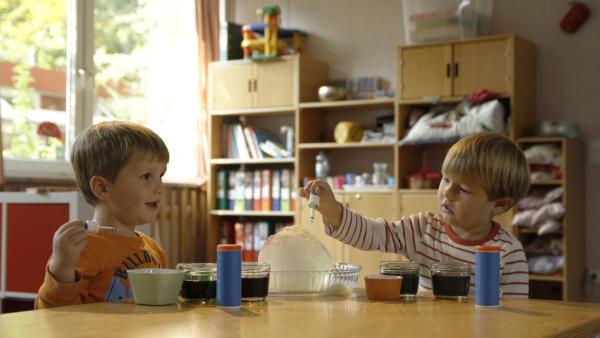 Zwei Kinder sitzen am Tisch. Vor ihnen ist eine Eiskugel und flüssige Farben und zwei Salzstreuer.