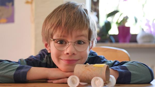Ein sechsjähriger Junge mit Brille sitzt lächelnd vor einem selbstgebastelten Papierrollen-Auto.