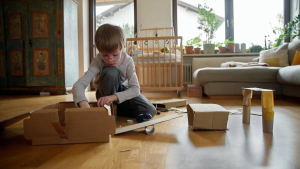 Ein Junge in grauer Kleidung kniet auf dem Wohnzimmerboden und bastelt mit Pappe eine Rennbahn.