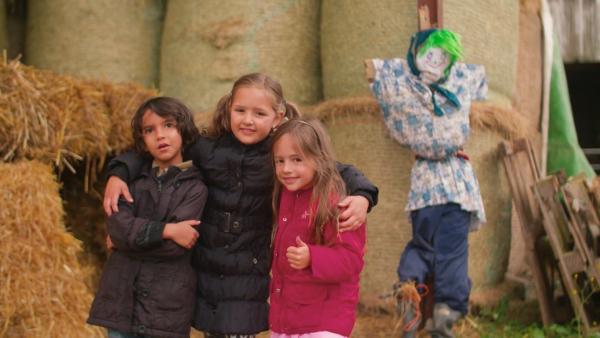 Drei Grundschulkinder in Winterjacken stehen vor einer selbstgebauten Vogelscheuche und Strohballen.