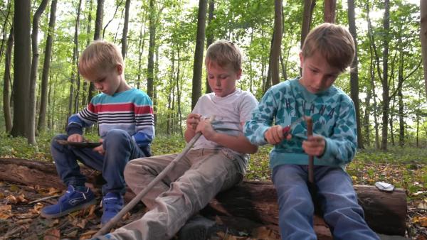 Drei Grundschulkinder sitzen auf einem Baumstamm und schnitzen mit Messern an Stöcken.