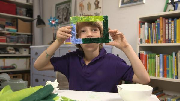 Eine Junge schaut durch seinen blau-grün bemalten Bilderrahmen und zeigt ihn stolz in die Kamera.