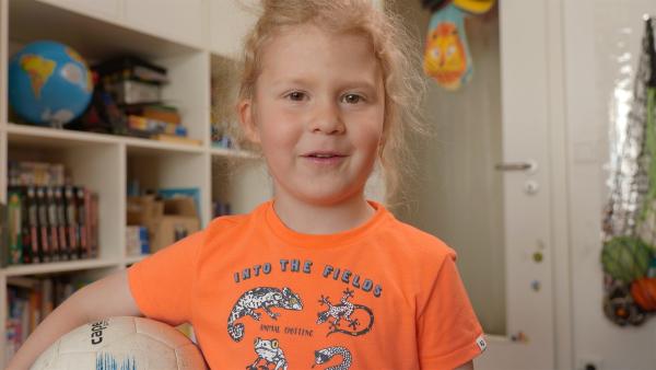 Ein Grundschulkind steht im Kinderzimmer. Es lächelt und hat einen Fußball unterm Arm.