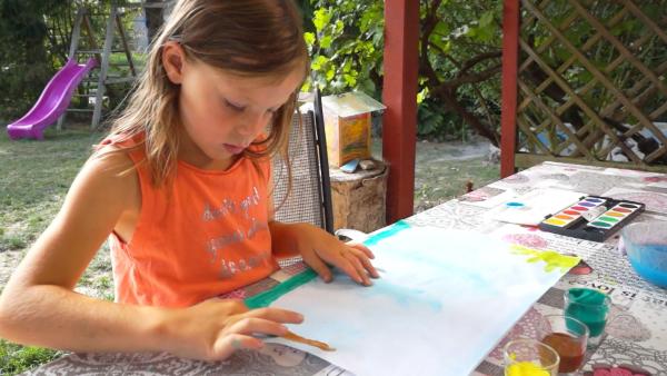 Ein Grundschulkind sitzt an einem Tisch im Garten und malt mit Fingermalfarben auf weißes Papier.