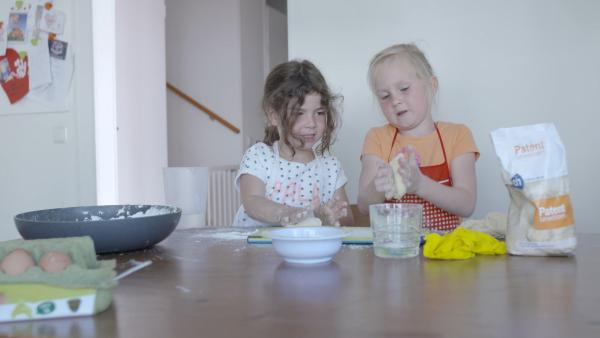 Zwei Mädchen sitzen an einem großen Tisch und kneten Teig. Neben ihnen stehen Materialien wie eine blaue Schüssel und eine Tüte Mehl.