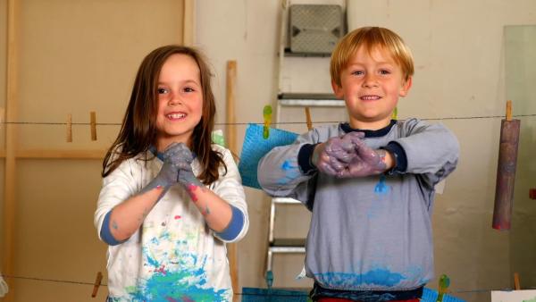 Zwei Vorschulkinder mit Händen voller bunter Farbe stehen vor einer Wäscheleine mit Bildern.