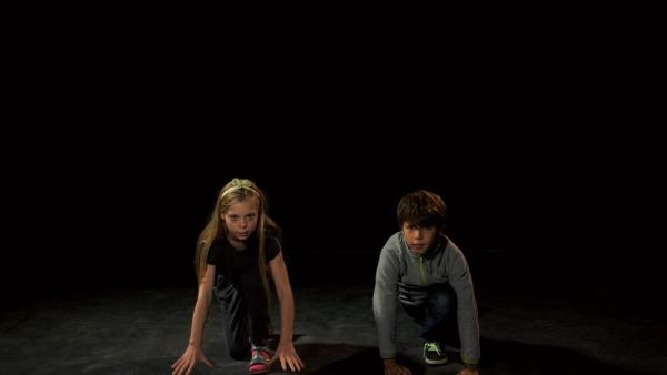 Ein Mädchen und ein Junge hocken vor einem schwarzen Hintergrund in Startposition auf dem Boden.