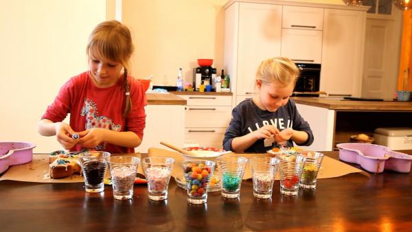 Zwei Mädchen stehen hinterm Tisch und schmücken ihren Kuchen mit vielen bunten Perlen.