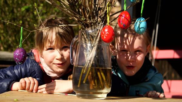 Zwei Mädchen schauen links und rechts hinter einem Strauß in einer Vase hervor. An dem Strauß hängen bunte Ostereier.