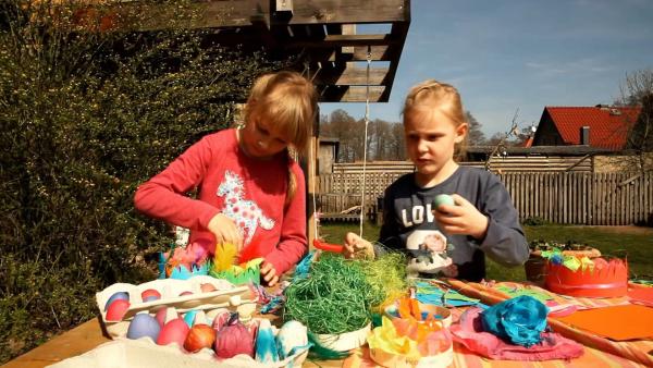 Zwei Mädchen sitzen im Garten hinterm Tisch. Sie füllen ihre Nester mit bunten Ostersachen.