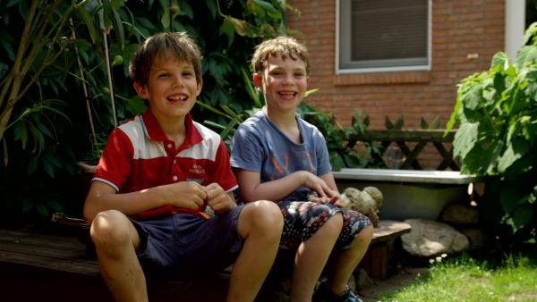 Zwei Jungs sitzen im Garten auf einer Holzbank und lachen in die Kamera.