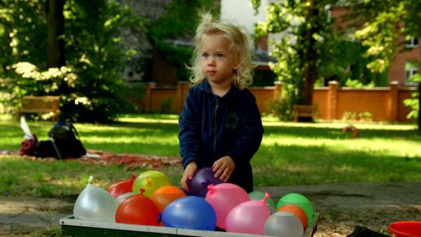 Ein Junge steht draußen vor einer Kiste vieler bunter Wasserballons. Er nimmt sich den lila Ballon.