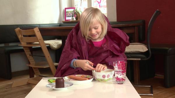 Ein Mädchen sitzt am Tisch und hat eine große, rote Jacke an. Sie isst aus einer Schüssel Suppe.