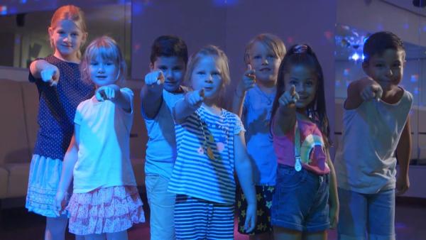 Sieben Kinder tanzen, lachen und strecken einen Arm nach vorne. Das Partylicht ist blau.