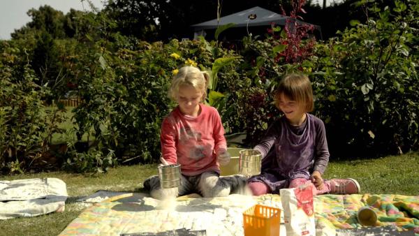Zwei Mädchen sitzen nebeneinander auf einer Decke in einem Garten und sieben Mehl auf Papier.