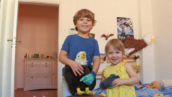 Ein Junge und eine Mädchen stehen in einem Kinderzimmer und lächeln. Der Junge hält einen Adler als Kuscheltier.
