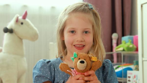 Ein Mädchen steht in einem Kinderzimmer und lächelt. Es hält einen Teddybären mit beiden Händen.