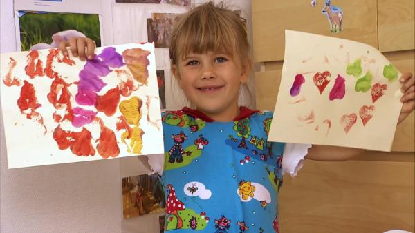 Ein Mädchen lächelt und zeigt ihr Bild, welches sie mit einem Stempel aus Kartoffeln bedruckt hat.