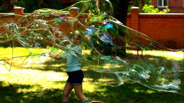 Ein Mädchen läuft bei Sonnenschein durch den Garten und zieht große Seifenblasen hinter sich her.