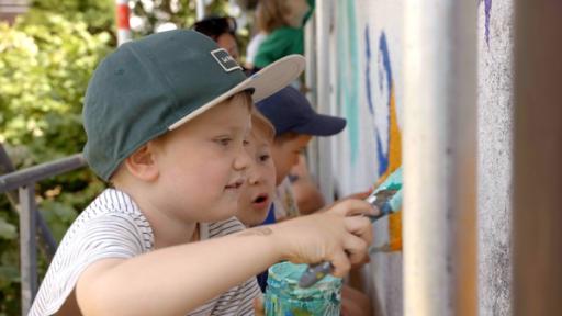 Ein Junge mit einer Kappe steht vor einer Mauer. Er hat ein Glas mit Farbe und einen Pinsel in der Hand. Er streicht die Farbe an die Wand. Im Hintergrund sind weitere Kinder zu sehen.