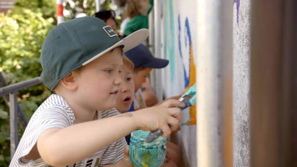 Ein Junge mit einer Kappe steht vor einer Mauer. Er hat ein Glas mit Farbe und einen Pinsel in der Hand. Er streicht die Farbe an die Wand. Im Hintergrund sind weitere Kinder zu sehen.