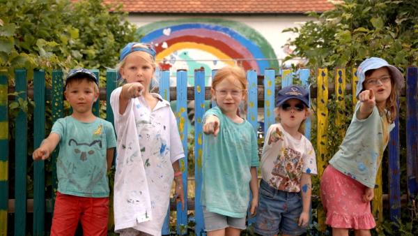 Fünf Kindergartenkinder stehen vor einer bemalten Hauswand. Sie zeigen mit den Fingern nach vorn.