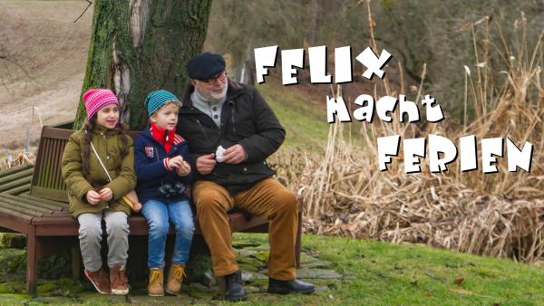 Felix sitzt mit einem Mädchen und seinem Opa auf einer Bank welche an einem Baum lehnt.