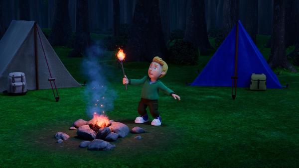 James steht neben einem Lagerfeuer auf einer Lichtung. Er schaut ängstlich auf eine brennende Fackel, die er in der Hand hält. Im Hintergrund links und rechts ein Zelt vor einem dunklen Wald.