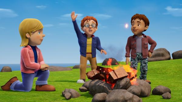 Zwei Jungs und ein Mädchen an einem Lagerfeuer, das von Steinen umgeben ist. Im Hintergrund das Meer.