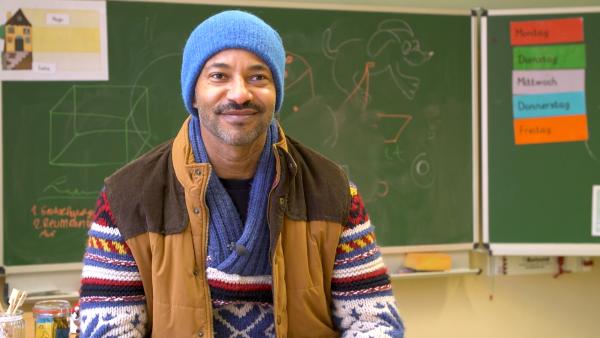 Tyron Ricketts sitzt in einem Klassenzimmer vor einer Tafel und lächelt.