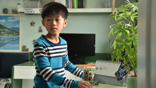 Der Junge Mo Ka-Keung steht an einem Schreibtisch. Darauf befindet sich ein Wochenkalender, im Hintergrund ein Bildschirm.