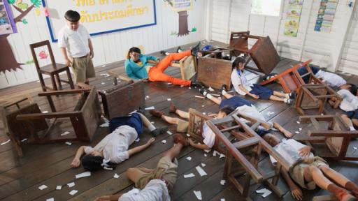 Rechte: SR/Thai PBS‘s Ein Junge steht in einem Klassenzimmer vor einer Tafel und schaut bedächtig nach unten. Neben ihm liegen Mitschüler bewusstlos am Boden.