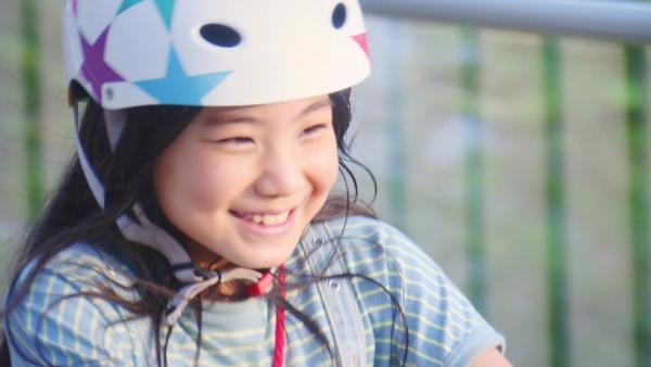 Fumis lächelnd und sehr nah. Sie trägt einen Fahrradhelm auf dem Kopf. Im Hintergrund ein Geländer.