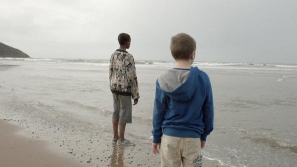 Zwei Jungen stehen am Meer. Sie sind rückseitig zu sehen. Bei trübem Himmel schauen sie zum Horizont.