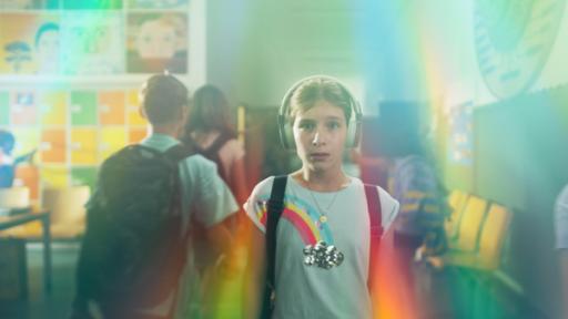 Eileen trägt Kopfhörer, ihren Ranzen und ein T-shirt mit Regenbogenmotiv